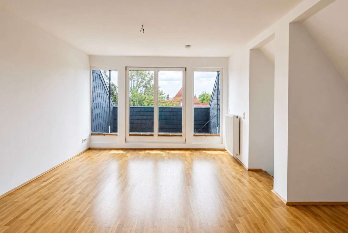 Bright, empty living room with large windows and a radiator on the right, overlooking a balcony with blue-tiled walls.