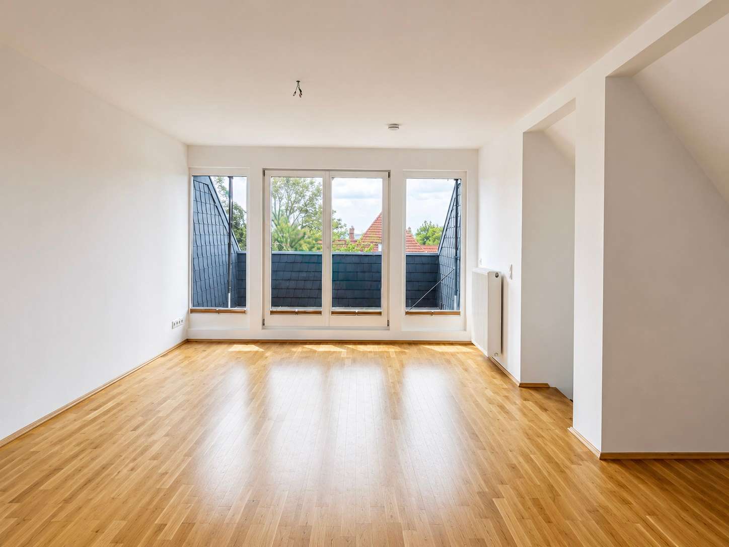 Bright, empty living room with large windows and a radiator on the right, overlooking a balcony with blue-tiled walls.