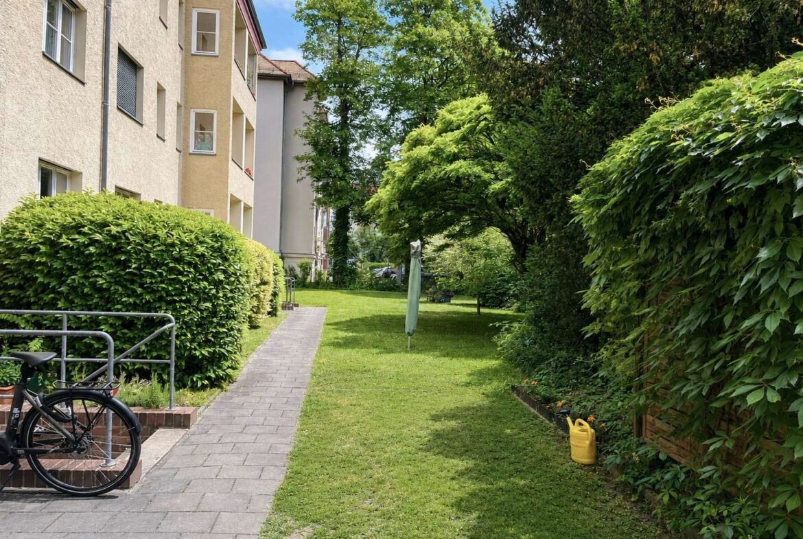 Residential courtyard scene with a paved path, a bicycle by a railing, and trimmed hedges along the building.