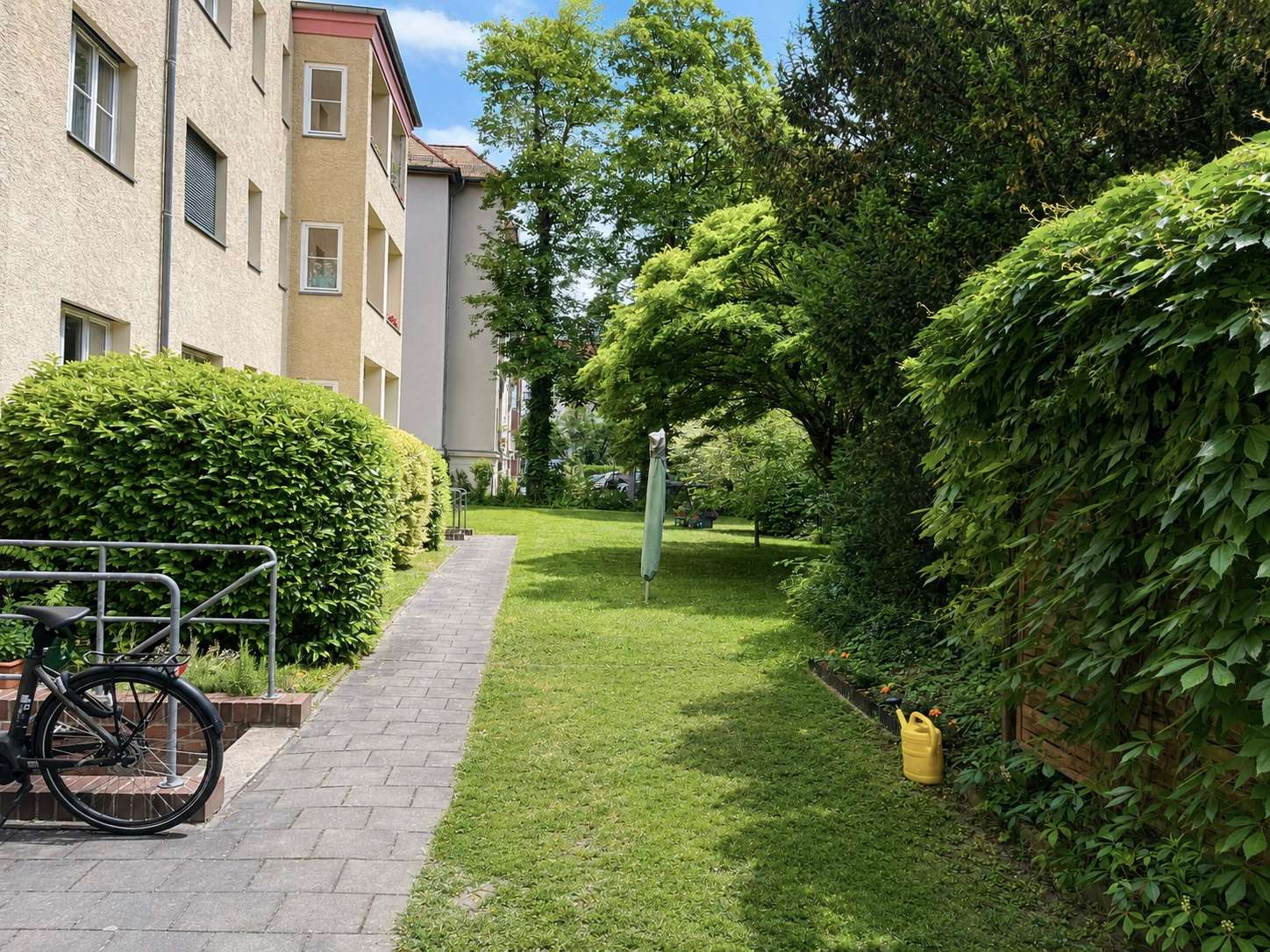 Residential courtyard scene with a paved path, a bicycle by a railing, and trimmed hedges along the building.