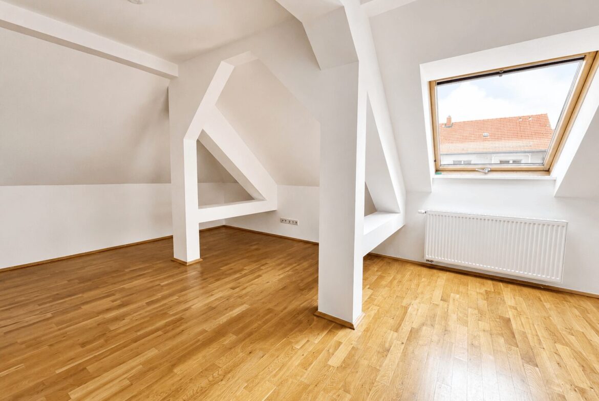 Bright attic room with white sloped ceilings, geometric beams, and warm wooden flooring; large skylight shows a red-tiled roof outside.