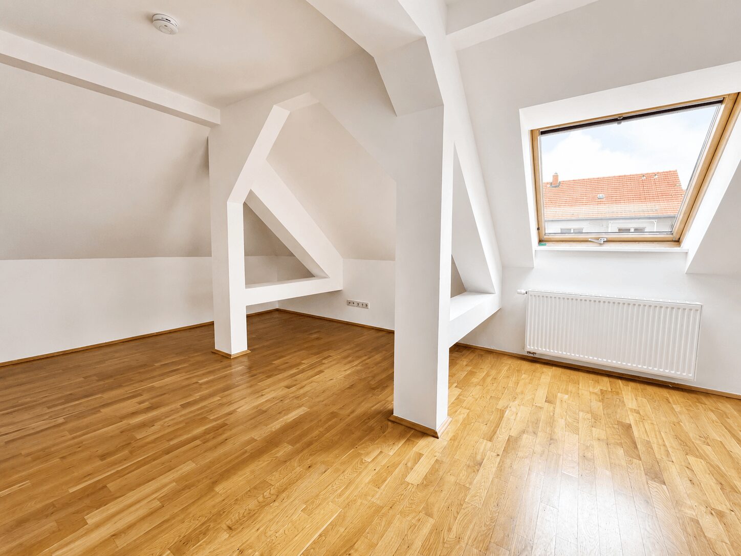 Bright attic room with white sloped ceilings, geometric beams, and warm wooden flooring; large skylight shows a red-tiled roof outside.
