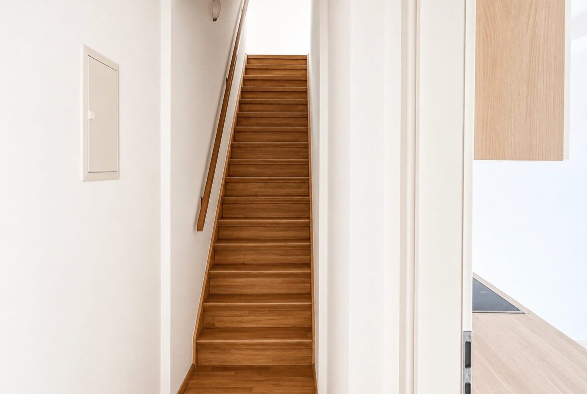 Wooden staircase in a bright white hallway with a wooden handrail on the left side.