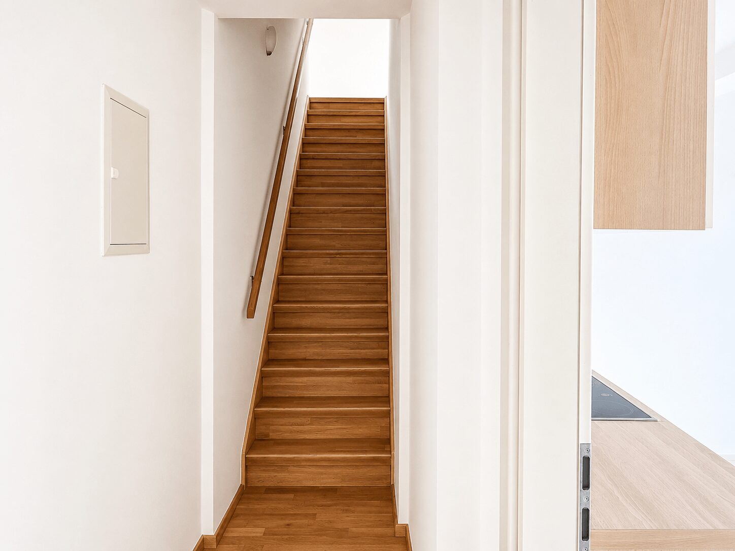 Wooden staircase in a bright white hallway with a wooden handrail on the left side.