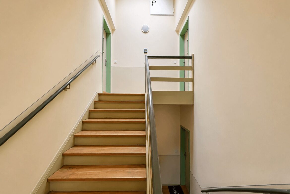 Indoor stairwell with beige walls, wooden steps, and gray metal handrails; green door frames on both sides and a upper landing railing.