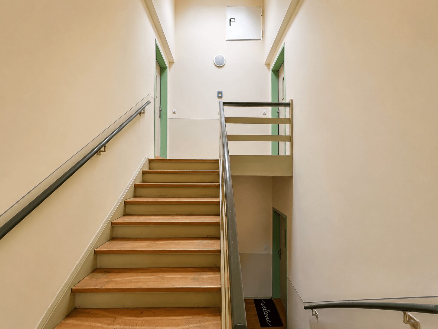 Indoor stairwell with beige walls, wooden steps, and gray metal handrails; green door frames on both sides and a upper landing railing.