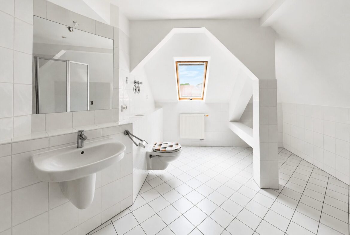 Bright white bathroom with tiled walls and floor, featuring a wall-mounted sink, mirrored cabinet, and wall-mounted toilet under a sloped ceiling with a skylight window.