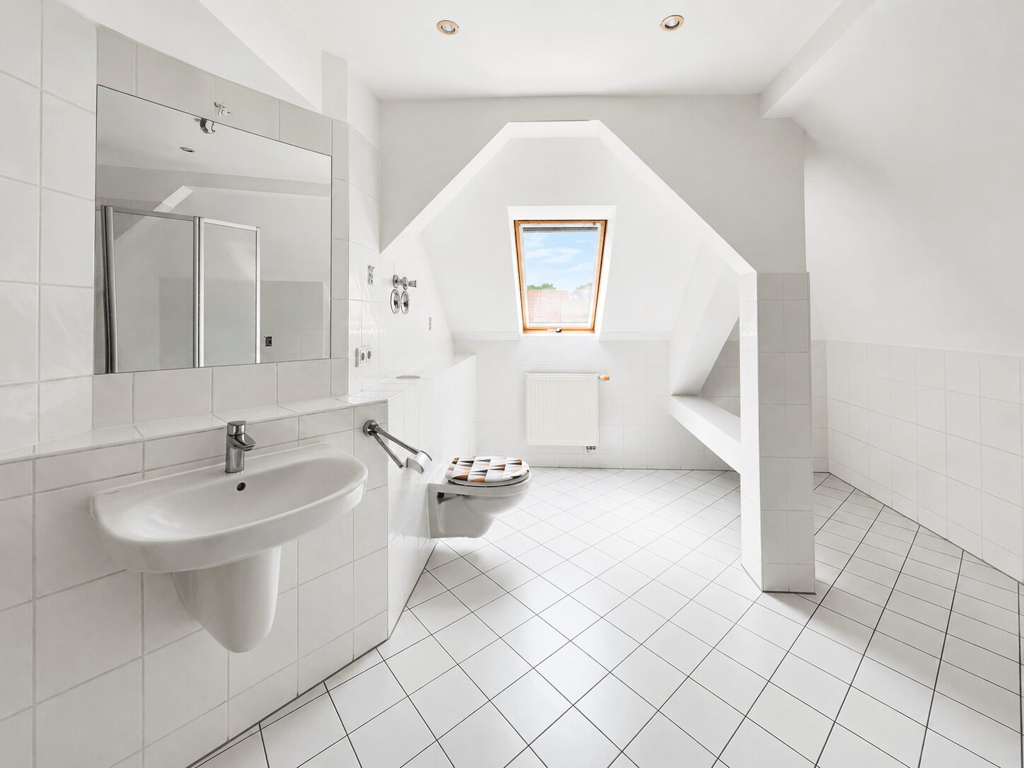 Bright white bathroom with tiled walls and floor, featuring a wall-mounted sink, mirrored cabinet, and wall-mounted toilet under a sloped ceiling with a skylight window.