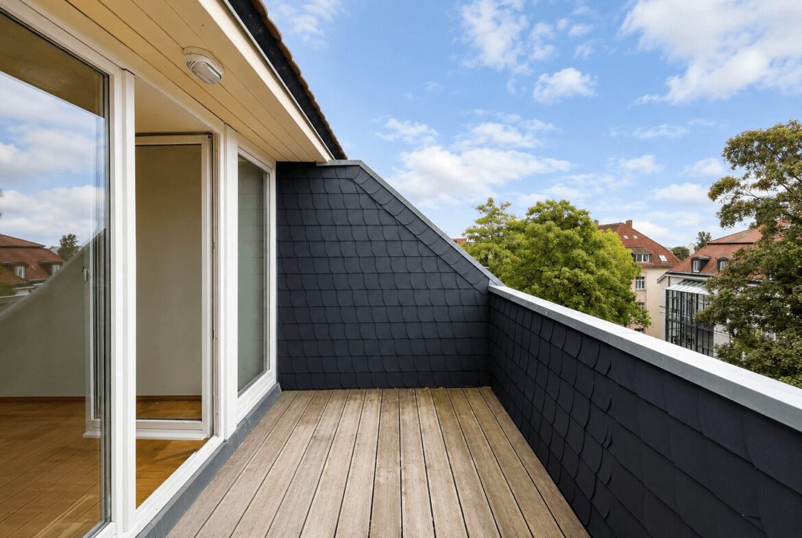 Wooden balcony deck with dark slate walls and glass sliding doors, looking out at trees and red-roofed buildings under a blue sky.