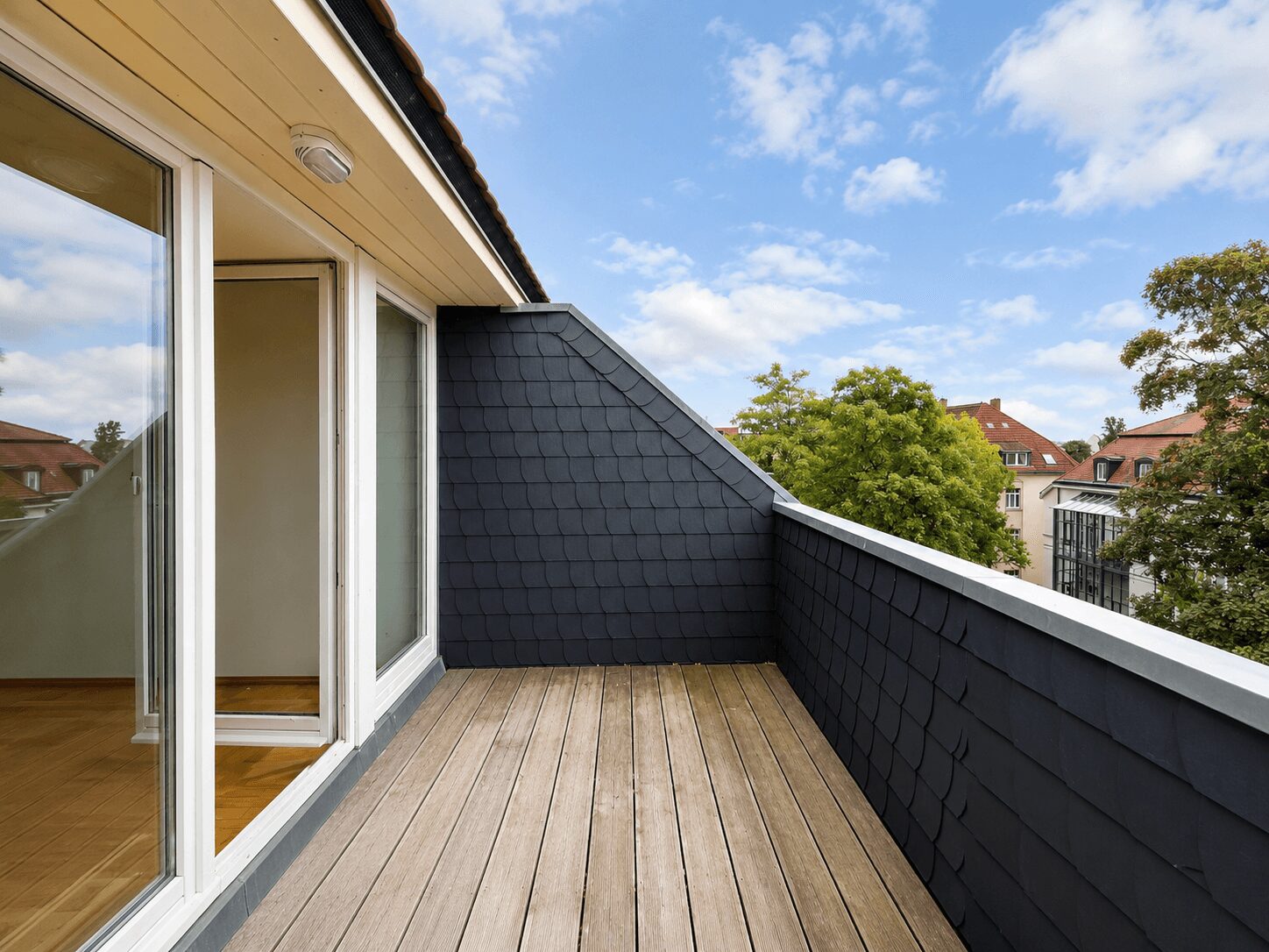 Wooden balcony deck with dark slate walls and glass sliding doors, looking out at trees and red-roofed buildings under a blue sky.