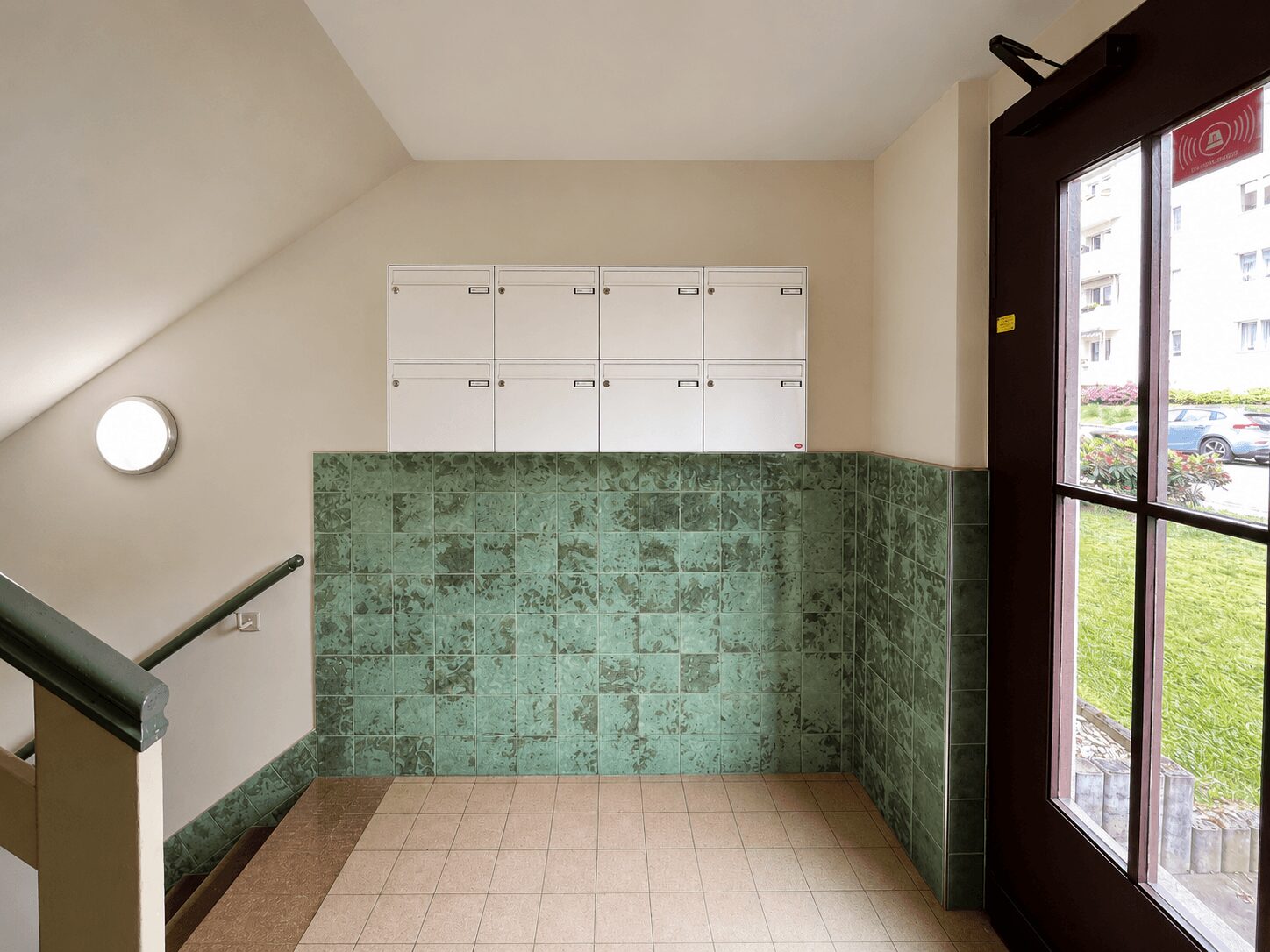 Stairwell landing with a row of white mailboxes above a green tiled wall and a glass door to the outside.