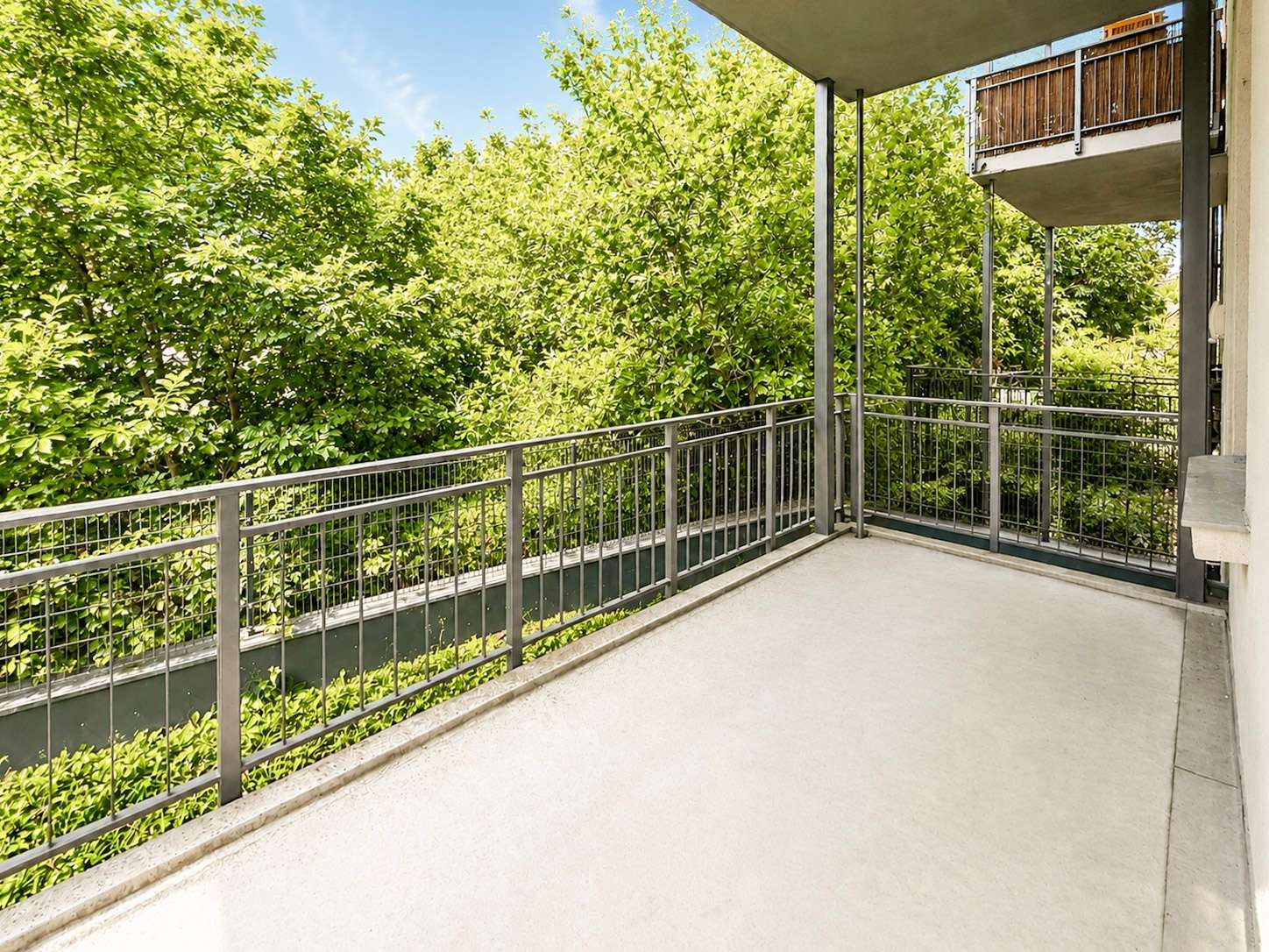 Concrete apartment balcony with a gray metal railing, overlooking dense green trees.