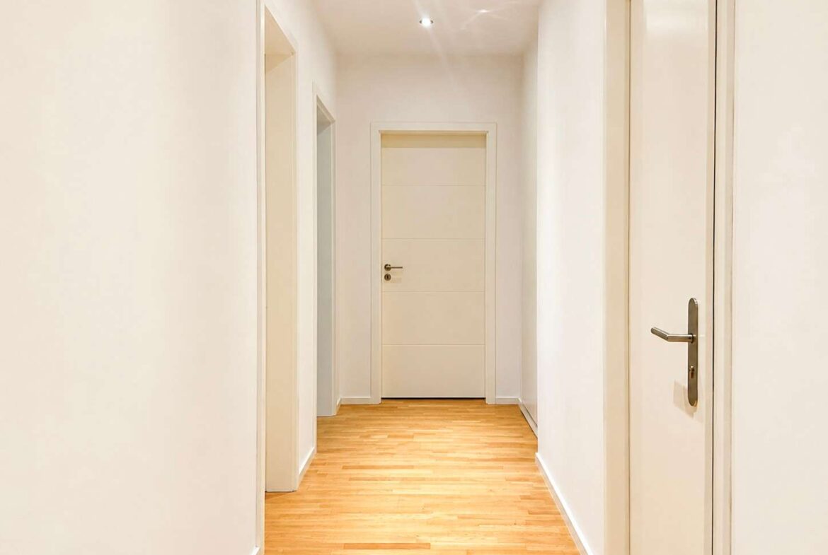 Bright white hallway with light wood flooring and several closed white doors along both sides, ending in a centered door at the far end.