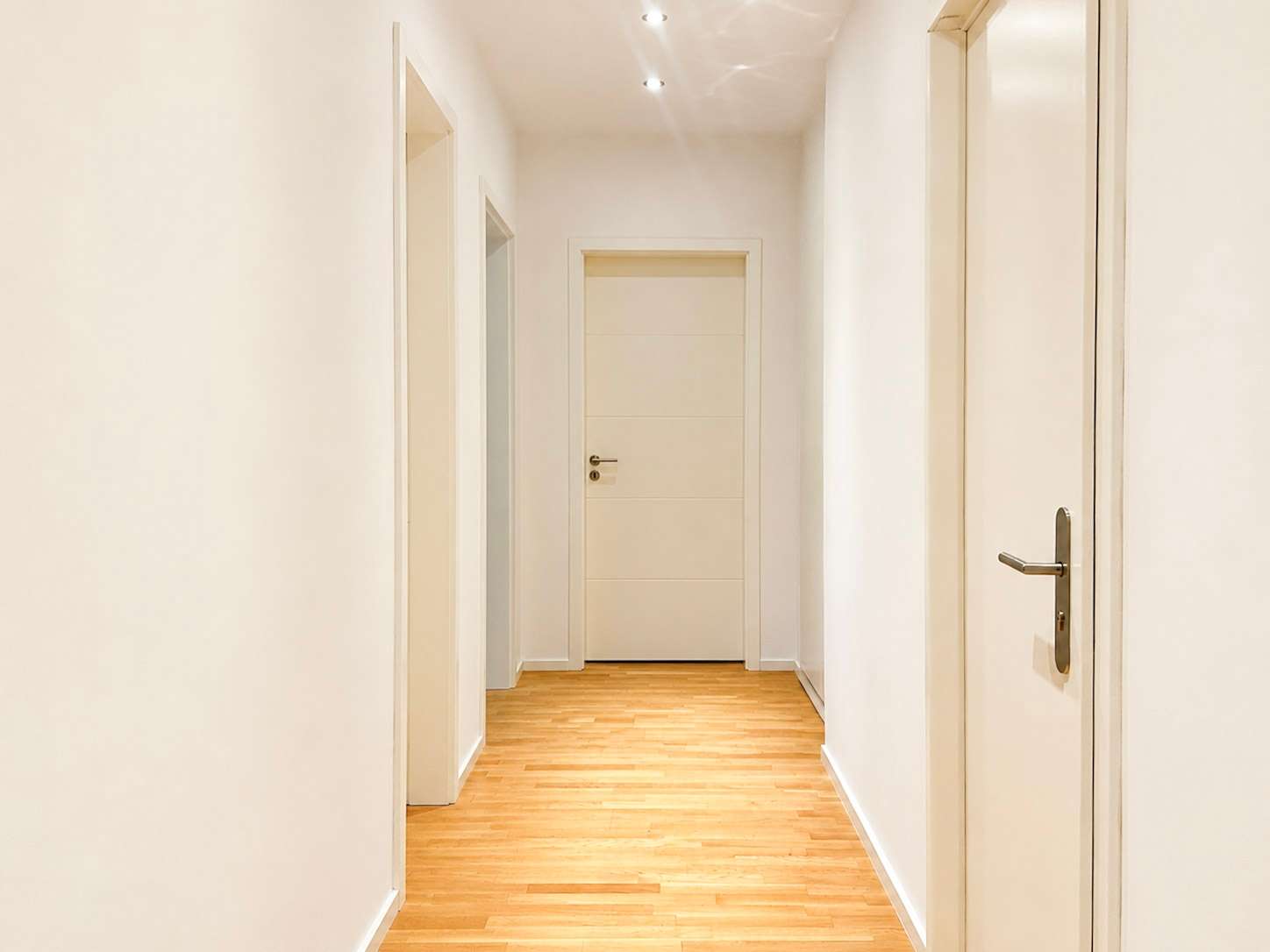 Bright white hallway with light wood flooring and several closed white doors along both sides, ending in a centered door at the far end.