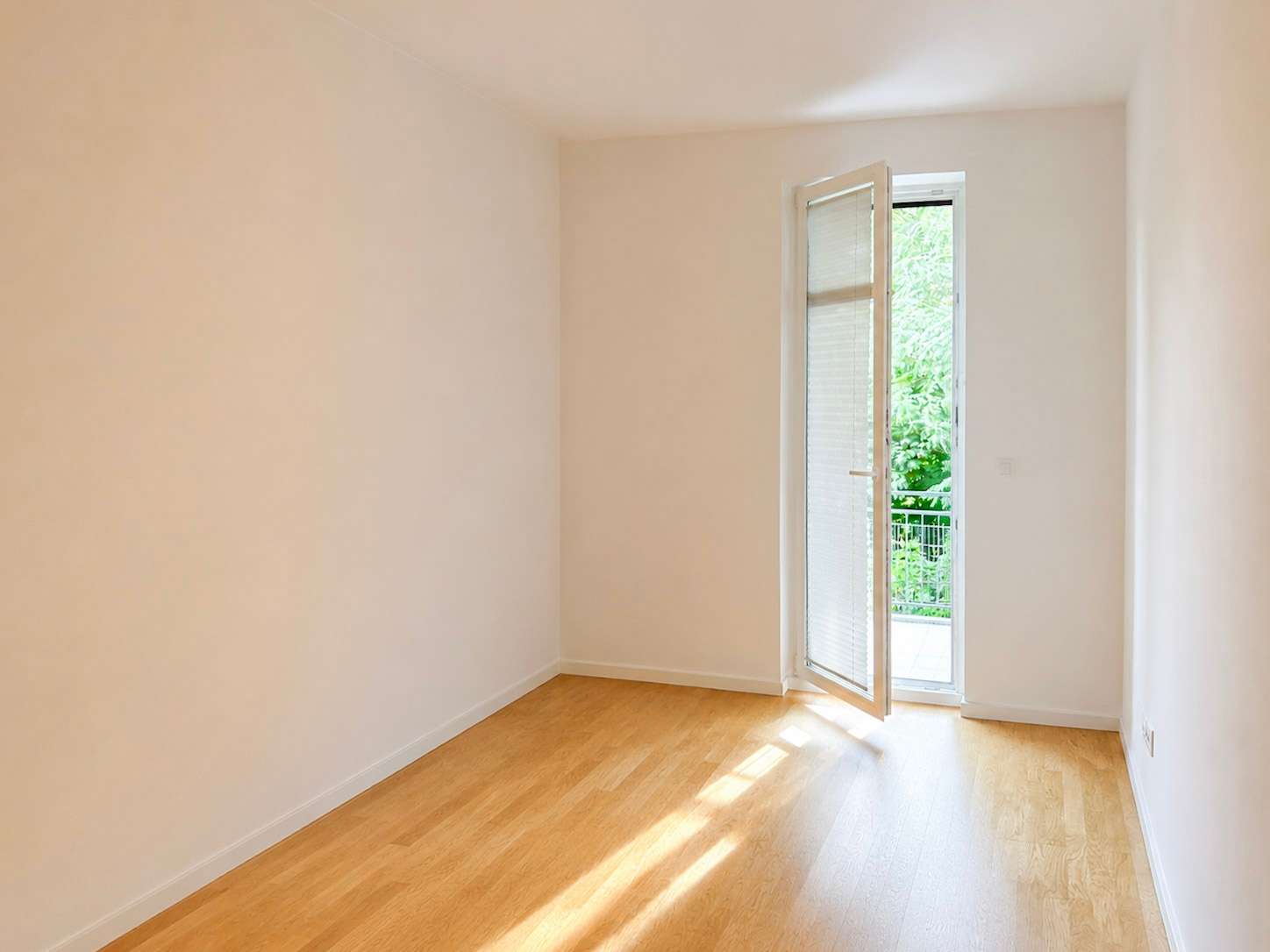 Bright empty room with light wood flooring and an open glass door leading to a green outdoor balcony area