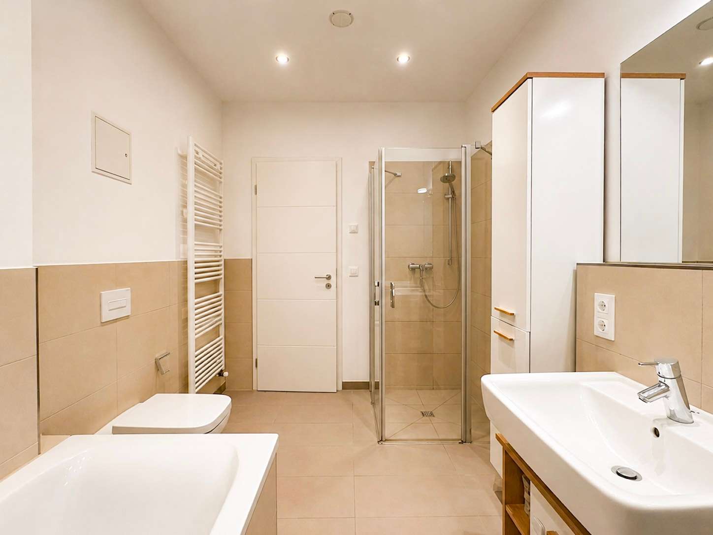 Bright modern bathroom with white fixtures: toilet, bidet, sink, glass shower, towel radiator and beige tiling.
