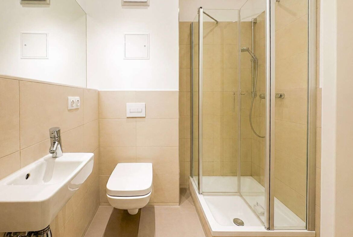 Modern beige-tiled bathroom with a wall-mounted toilet, rectangular sink, and a glass shower enclosure with chrome fixtures.