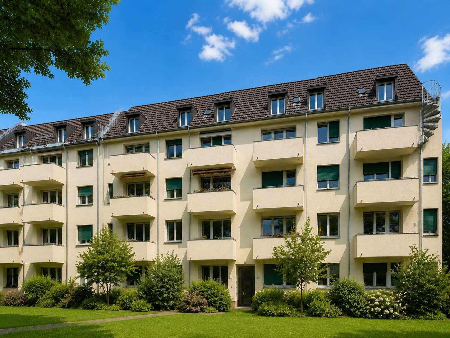 Three-story beige apartment building with balconies facing a green lawn, under a blue sky with a few clouds.
