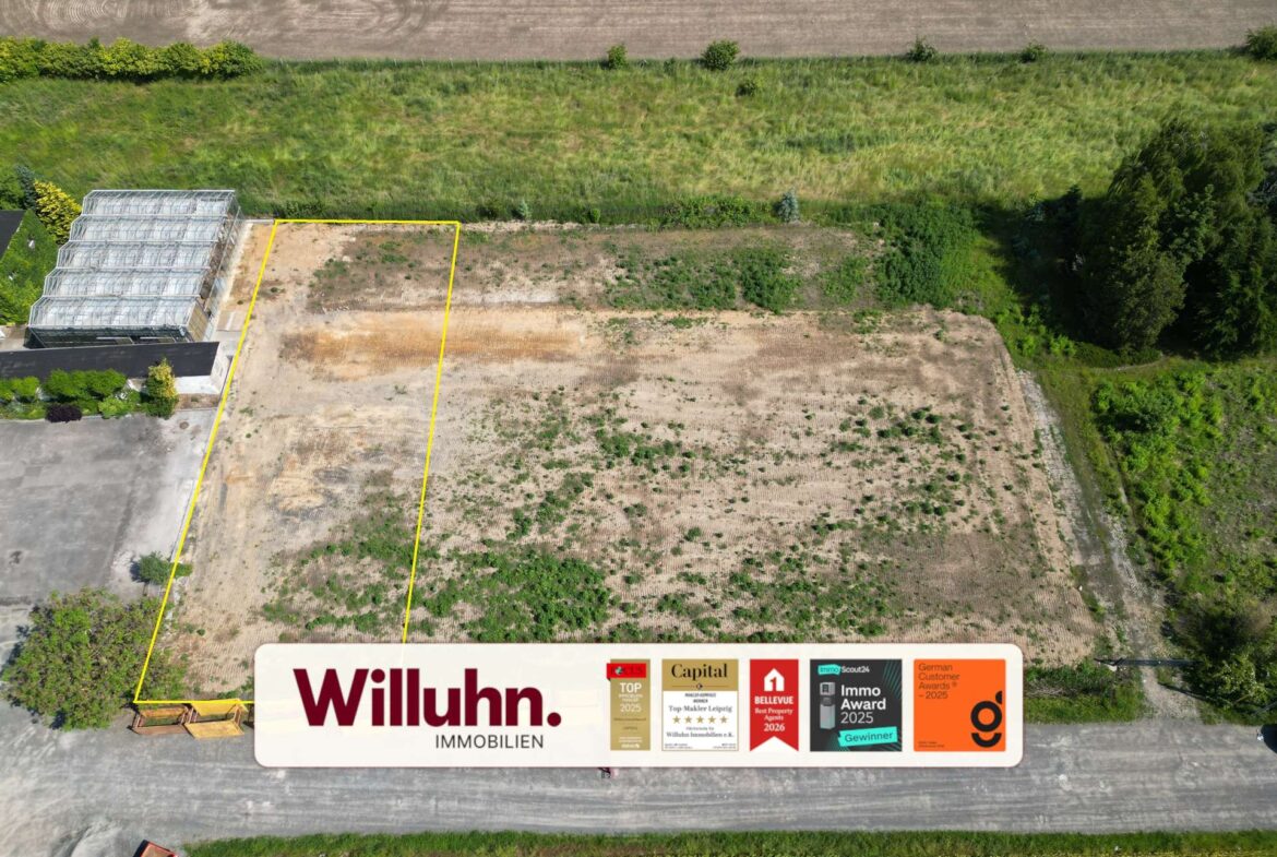 Aerial view of a vacant lot outlined in yellow beside a greenhouse, with a large Willuhn Immobilien sign at the edge of the frame.