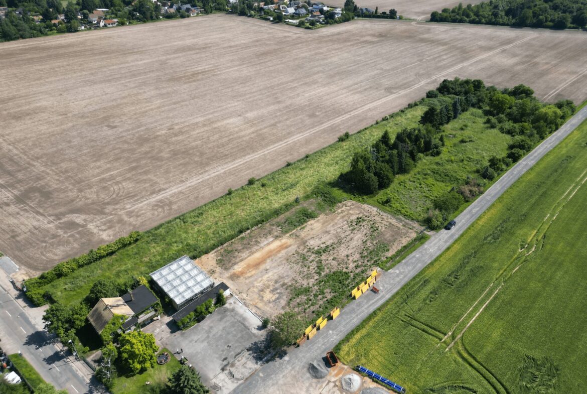Aerial view of a large harvested field bordered by a road, with a small village and trees in the distance.