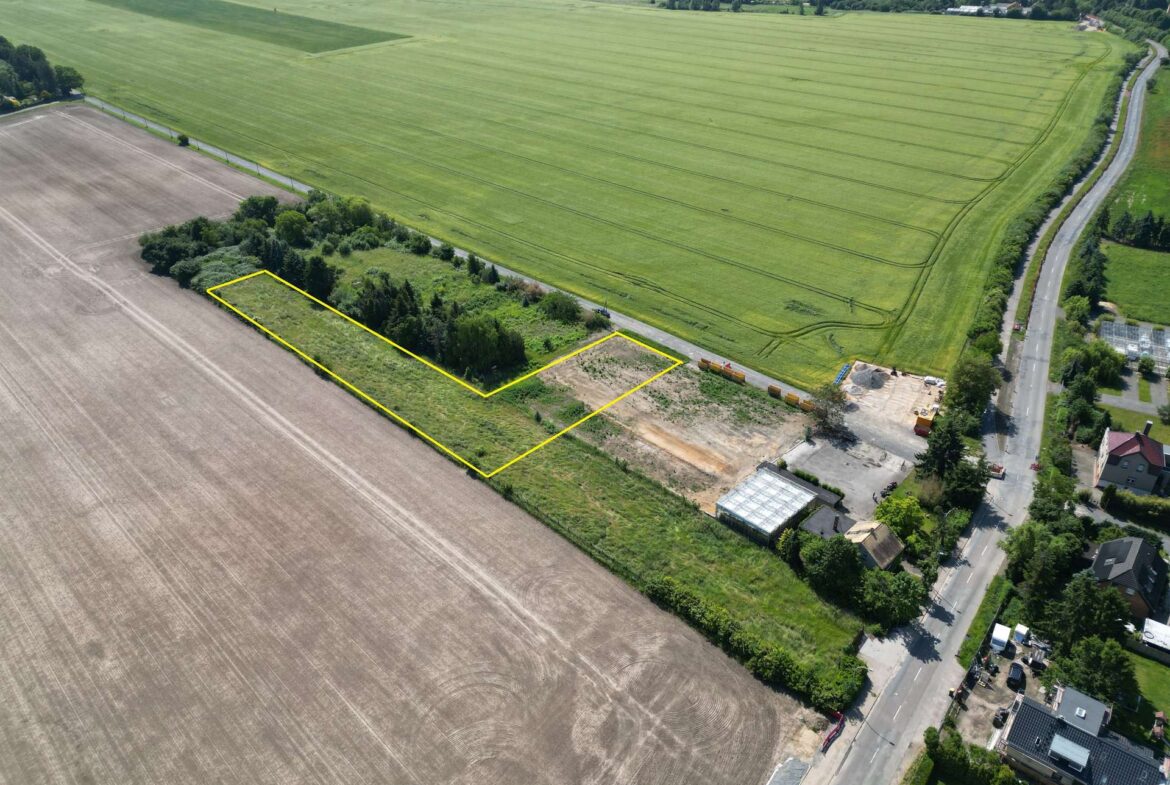 Aerial view of fields with two adjacent rectangular plots outlined in yellow near a road, showing construction equipment and a small building area nearby.