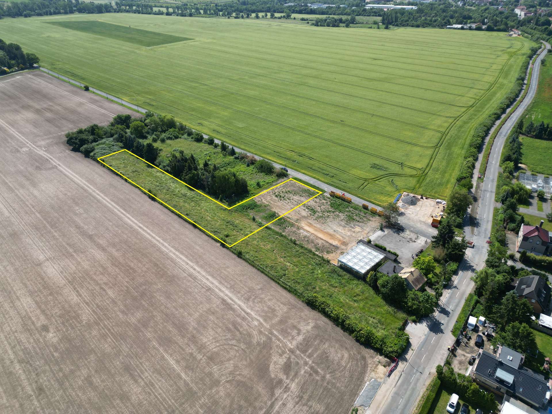 Aerial view of fields with two adjacent rectangular plots outlined in yellow near a road, showing construction equipment and a small building area nearby.