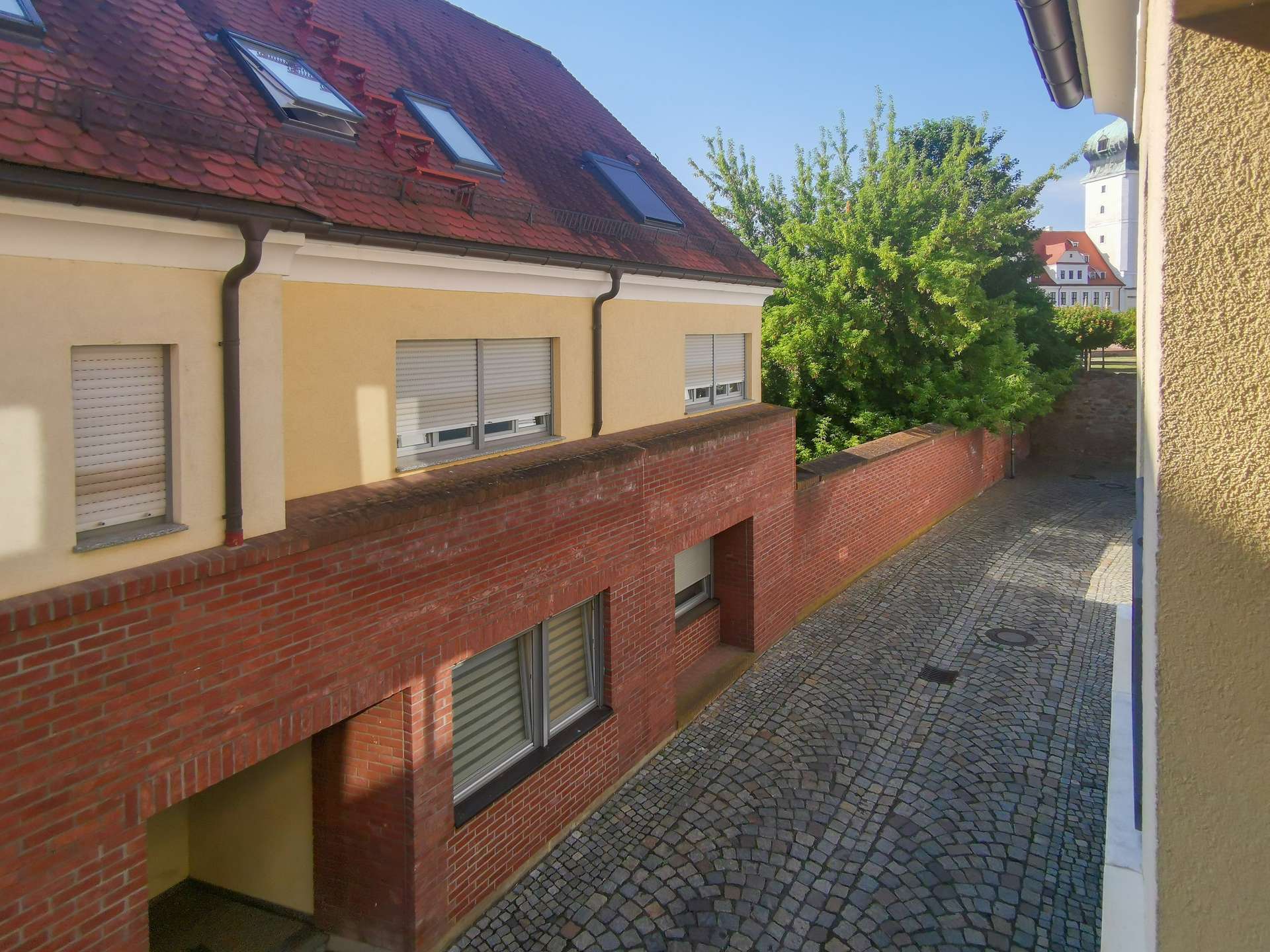 Narrow cobblestone street flanked by a beige building with a red brick base and red tile roof, with a green tree and a distant church tower in the background.