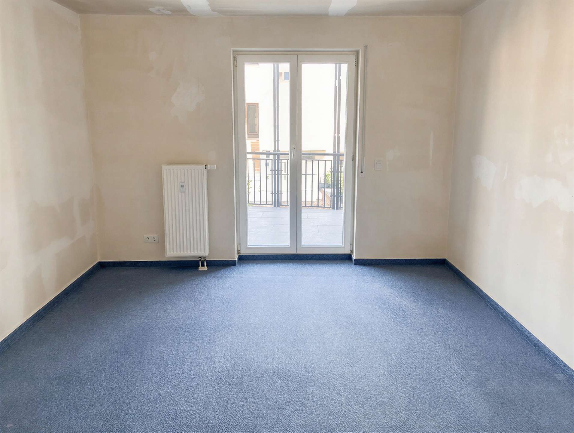 Empty, unfurnished room with blue carpet, a wall-mounted radiator on the left, and a glass door leading to a balcony.