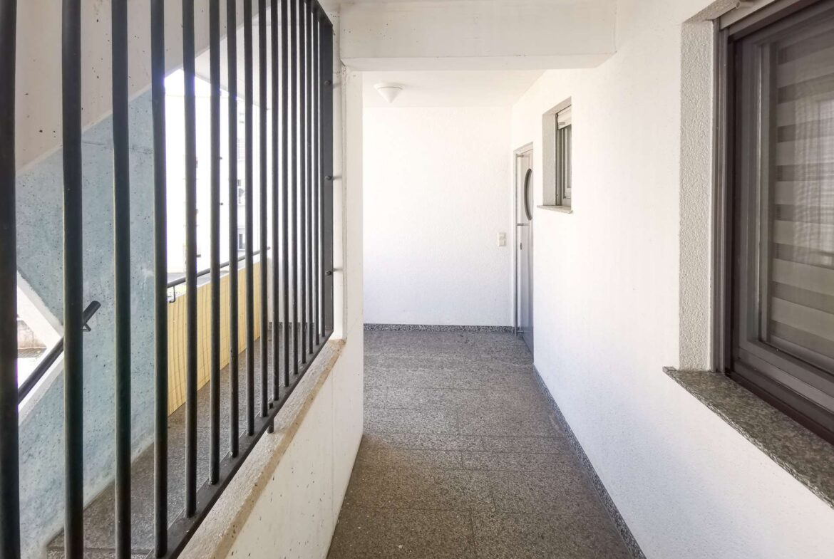 Narrow outdoor corridor with black vertical railing on the left, white walls, and a door at the far end.