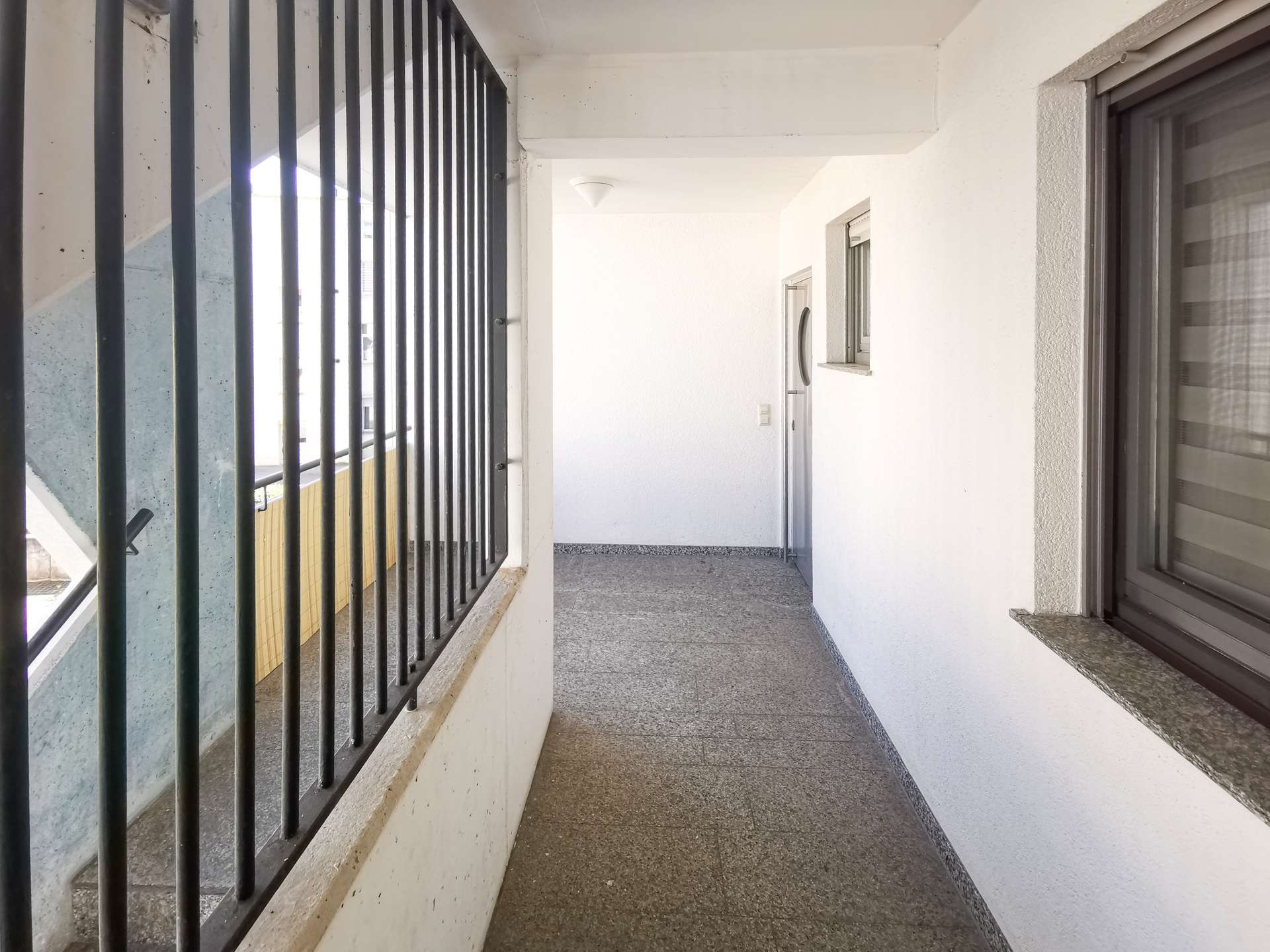 Narrow outdoor corridor with black vertical railing on the left, white walls, and a door at the far end.