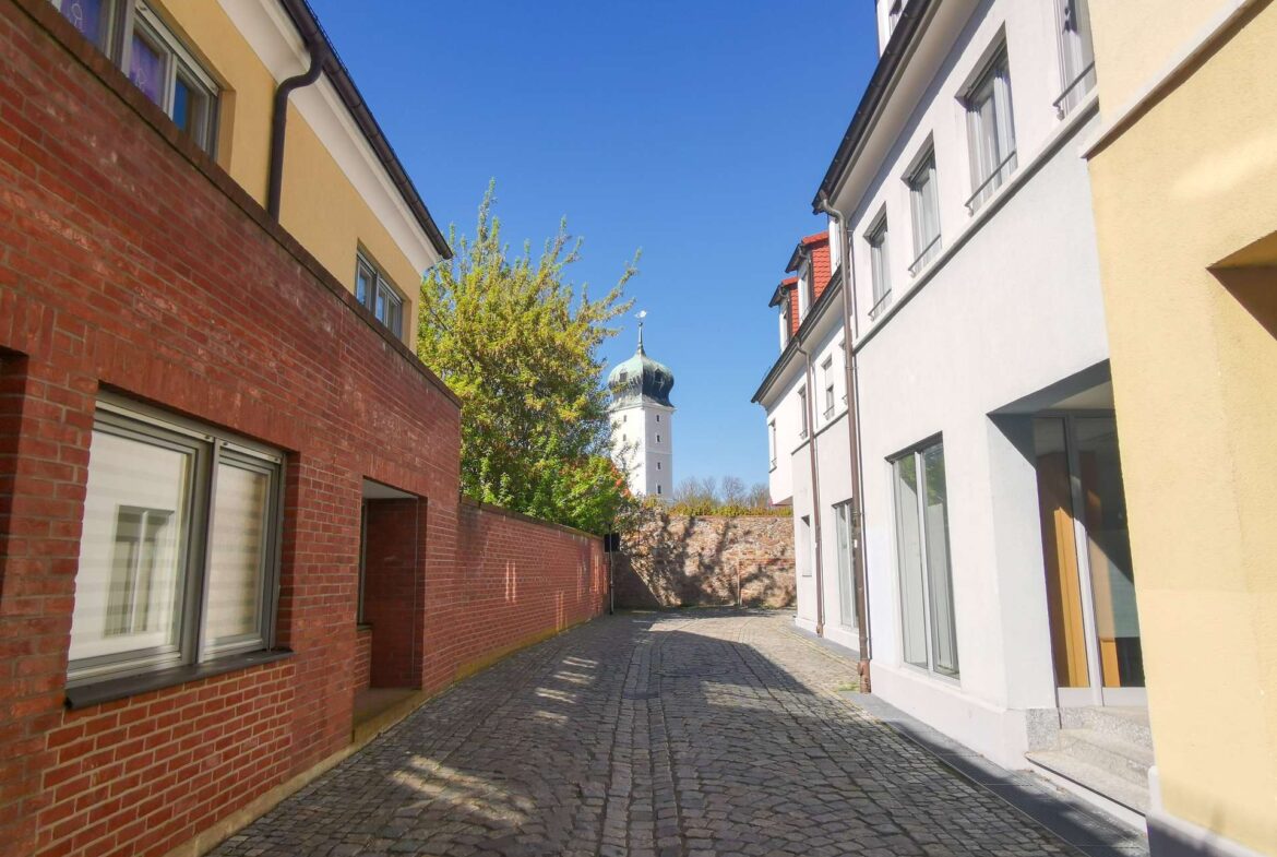 Narrow cobblestone alley between red brick left wall and light-colored right building, blue sky and a distant church tower with a green dome.