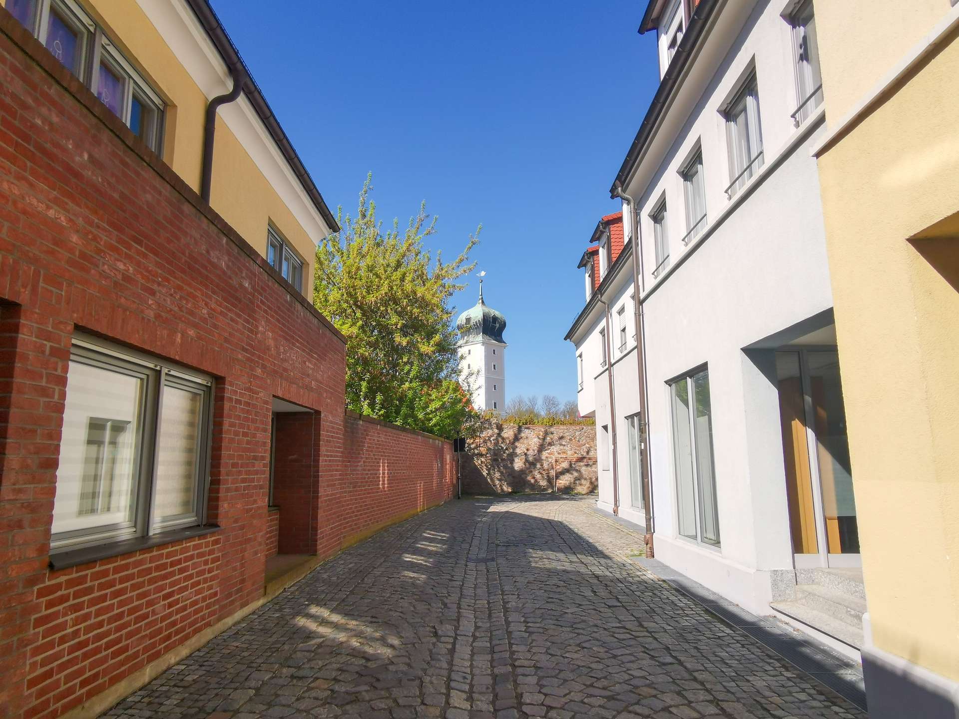 Narrow cobblestone alley between red brick left wall and light-colored right building, blue sky and a distant church tower with a green dome.