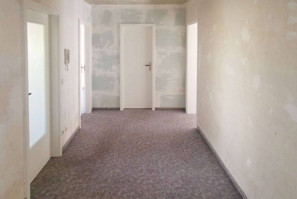 Empty hallway in a building under renovation; white doors along pale, patchy walls at the ends and sides.