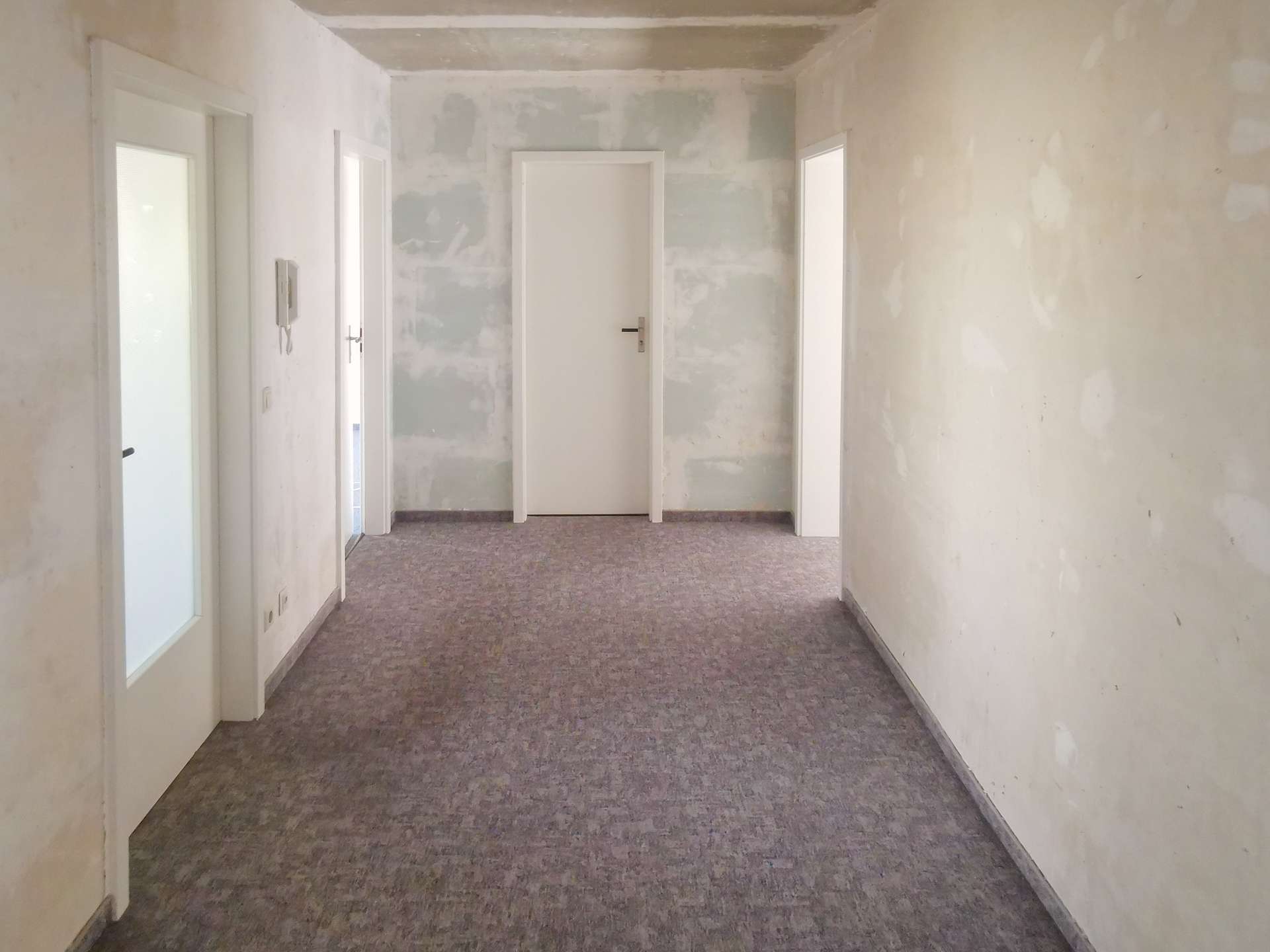 Empty hallway in a building under renovation; white doors along pale, patchy walls at the ends and sides.