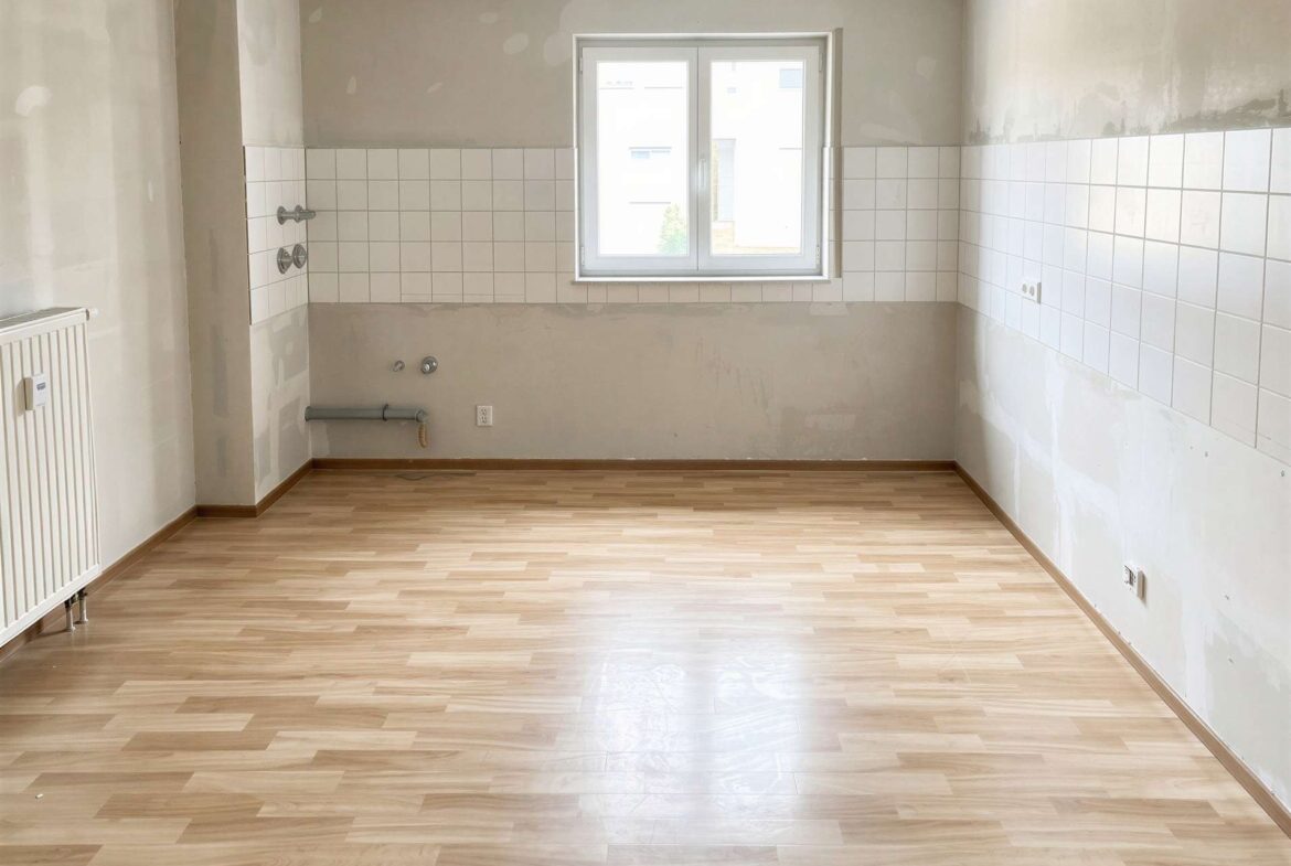 Empty kitchen awaiting installation: white tile splashback, bare walls, exposed pipes by the plumbing, and a centered window over light wooden flooring