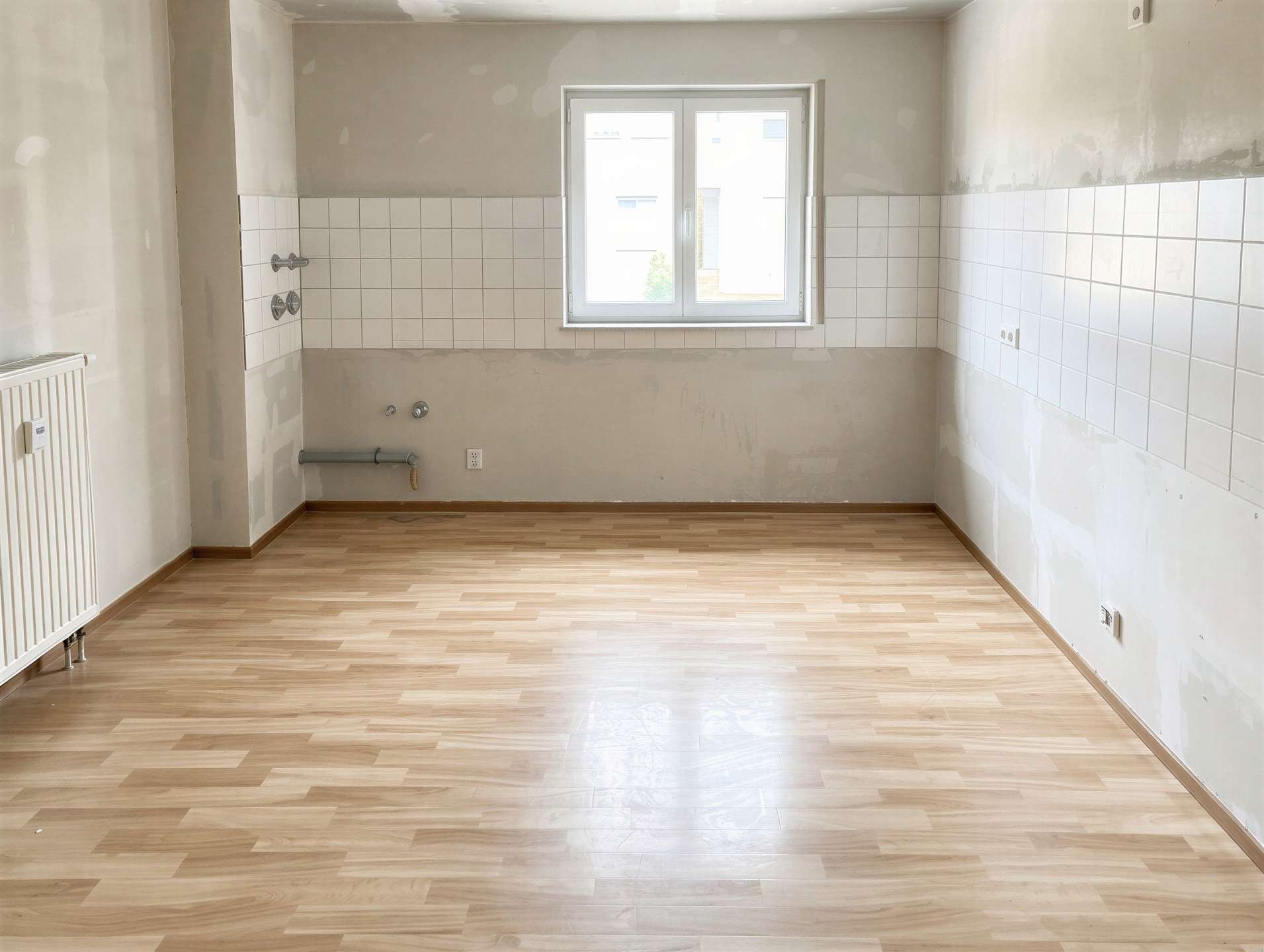 Empty kitchen awaiting installation: white tile splashback, bare walls, exposed pipes by the plumbing, and a centered window over light wooden flooring