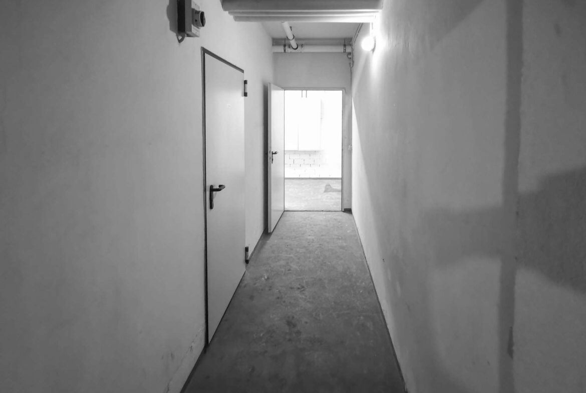Dim, narrow hallway with closed door on left and an open doorway at the far end leading to a bright room; exposed pipes along the ceiling.