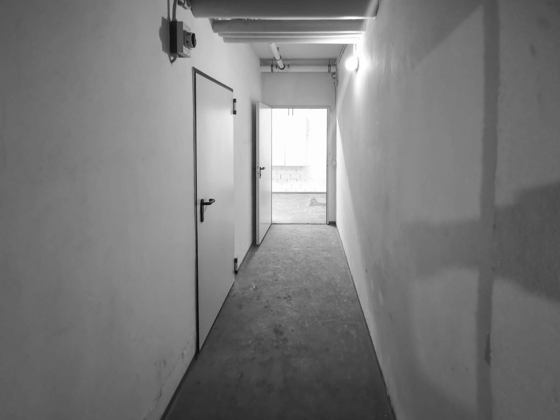 Dim, narrow hallway with closed door on left and an open doorway at the far end leading to a bright room; exposed pipes along the ceiling.