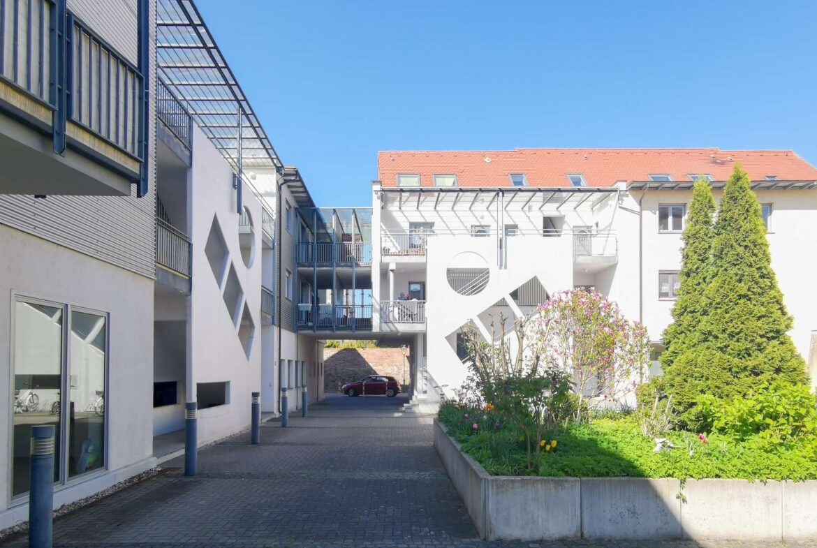 Sunny residential courtyard between white buildings with metal balconies and a paved walkway under a clear blue sky.
