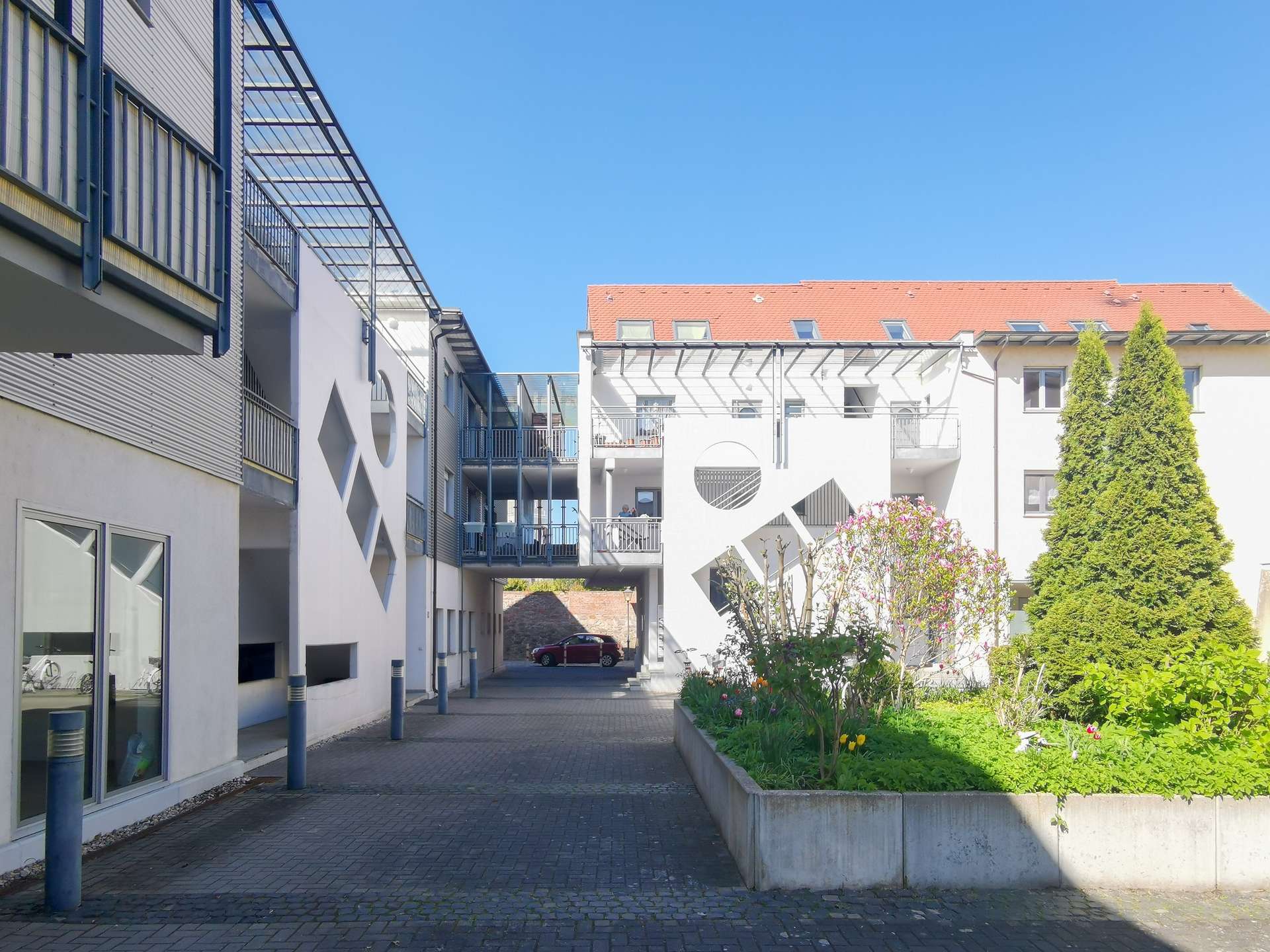 Sunny residential courtyard between white buildings with metal balconies and a paved walkway under a clear blue sky.
