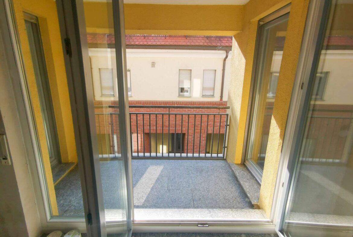 Open sliding doors lead to a small yellow-walled balcony with a metal railing overlooking a neighboring brick building, with paint cans on the left floor.
