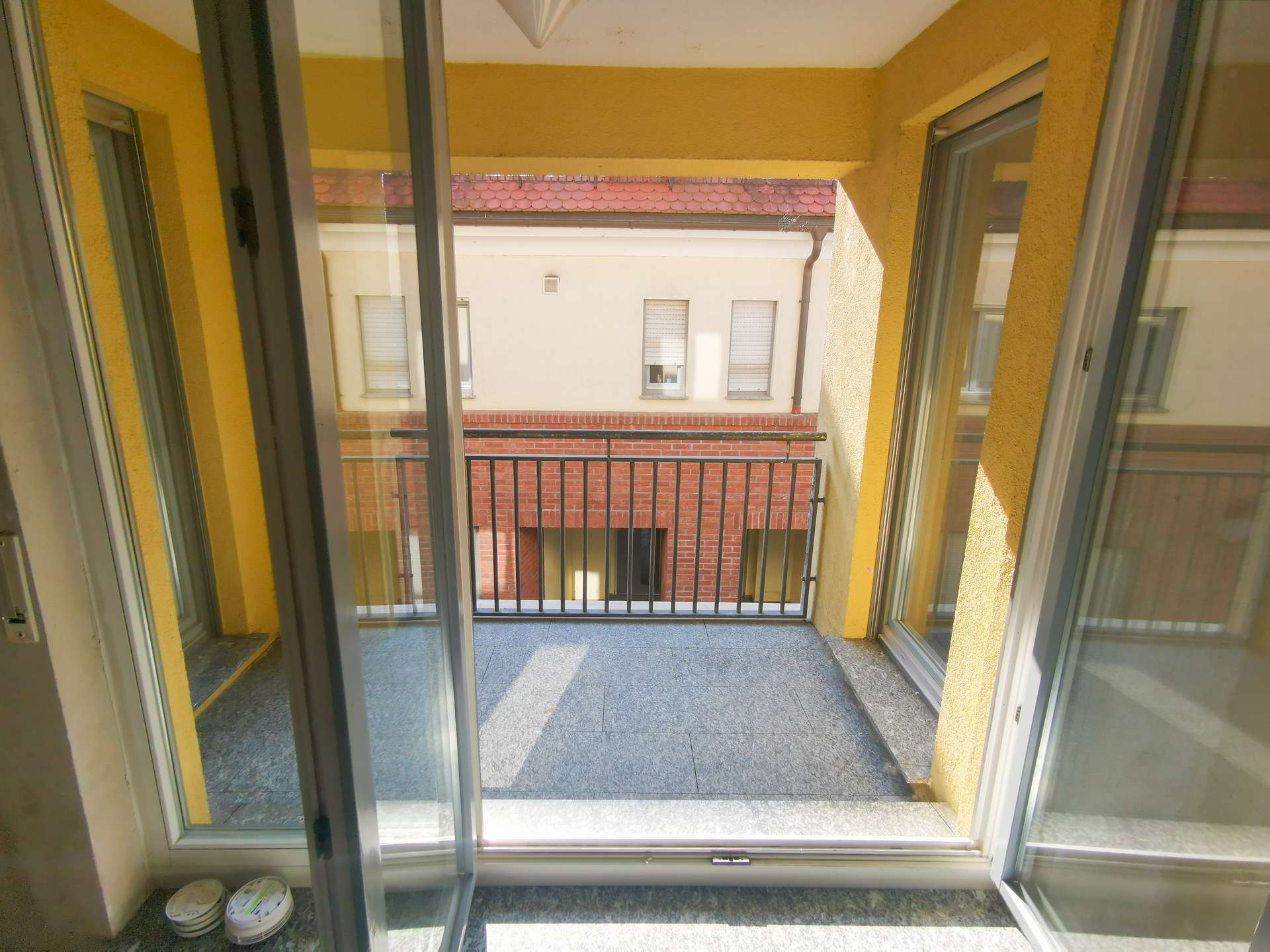 Open sliding doors lead to a small yellow-walled balcony with a metal railing overlooking a neighboring brick building, with paint cans on the left floor.