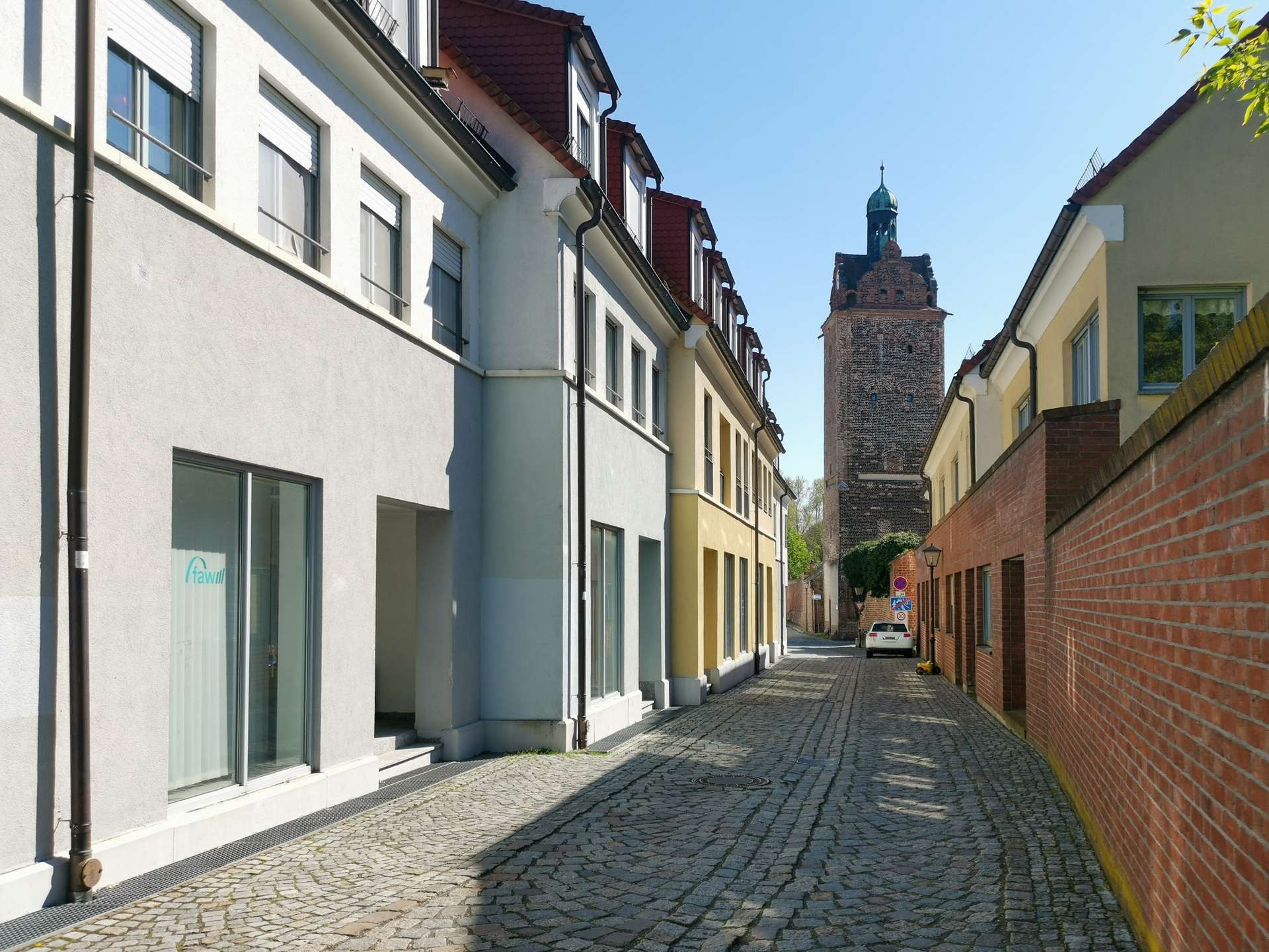 Narrow cobblestone street between pastel townhouses, with a historic brick tower visible in the distance under a blue sky.