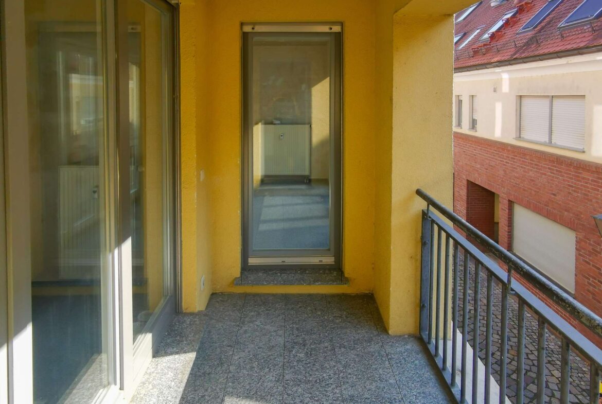 Yellow-walled apartment balcony with sliding glass doors, gray tiled floor, and a metal railing overlooking a neighboring red-brick building.
