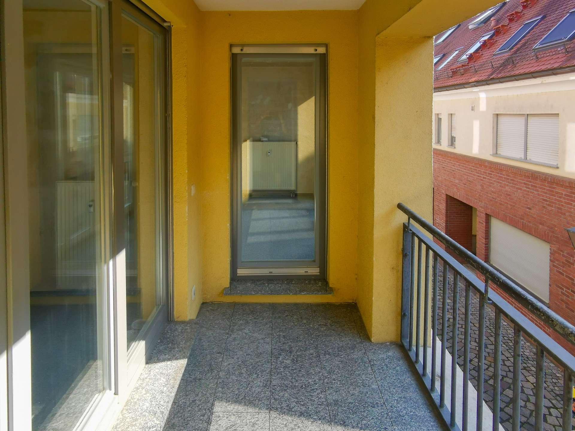Yellow-walled apartment balcony with sliding glass doors, gray tiled floor, and a metal railing overlooking a neighboring red-brick building.