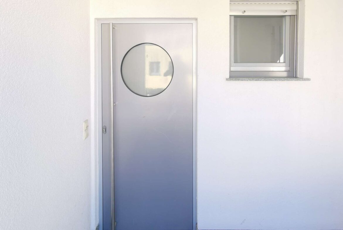 Grey metal exterior door with a circular window, beside a small rectangular window set in a white wall.