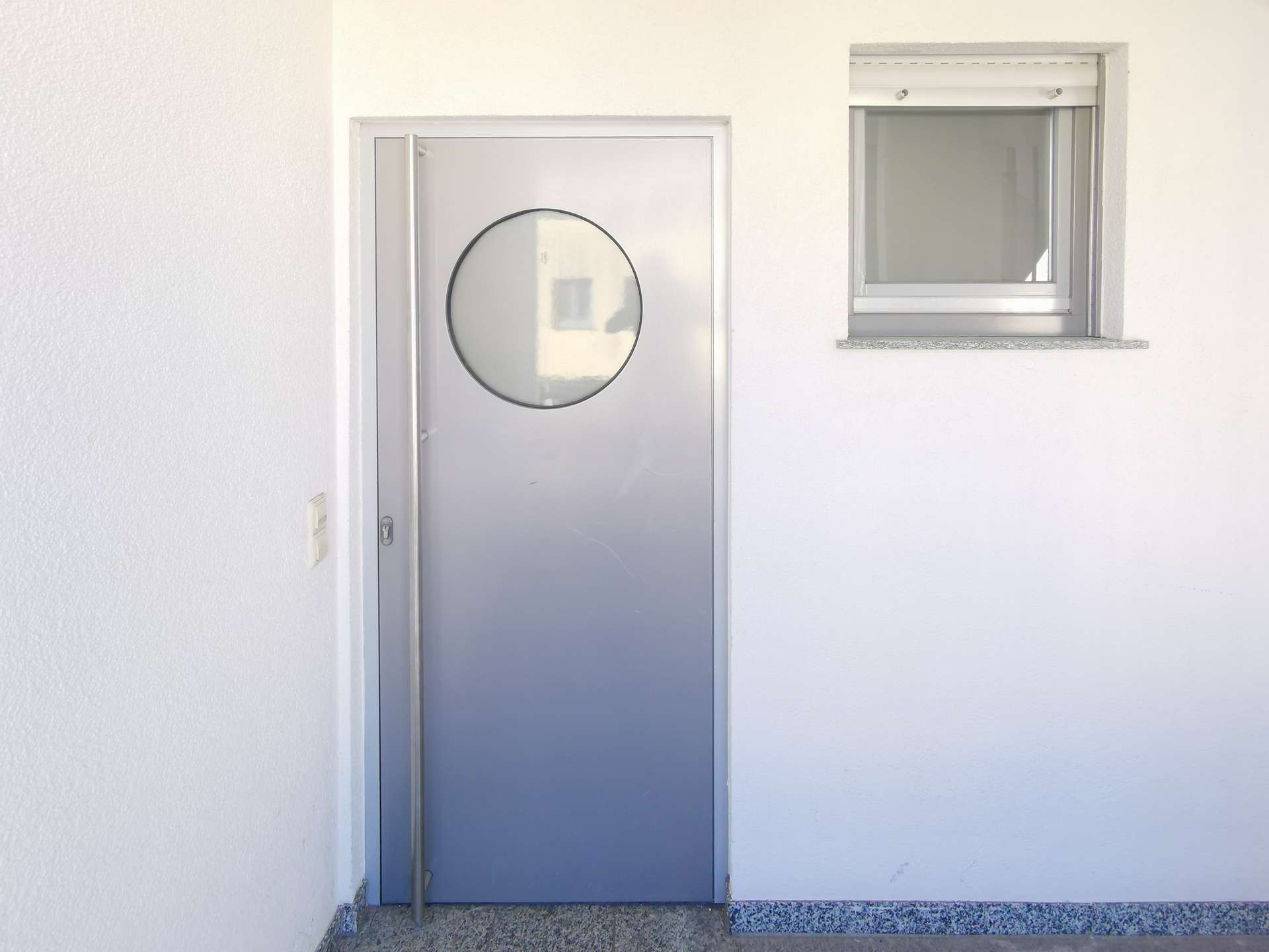 Grey metal exterior door with a circular window, beside a small rectangular window set in a white wall.