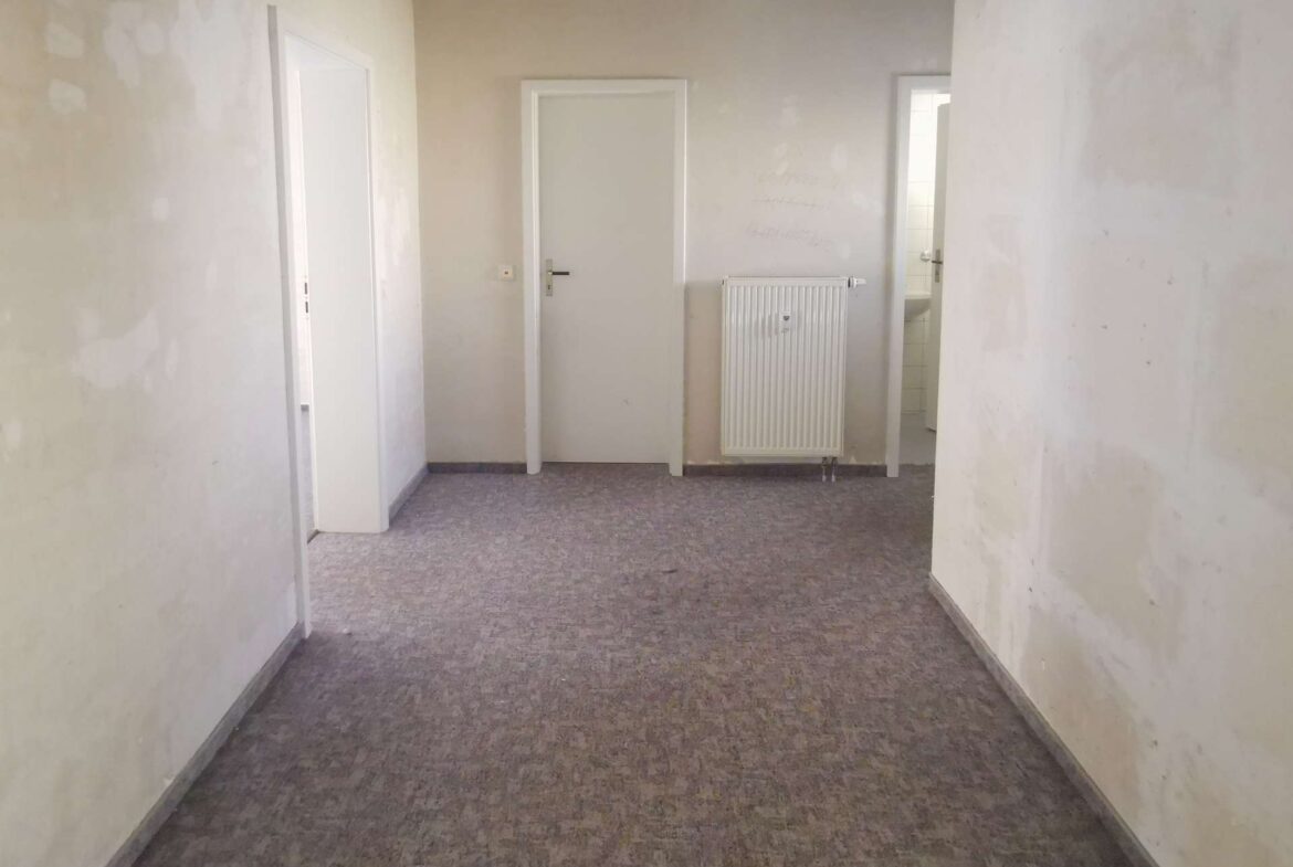 Unfinished indoor hallway with patched pale walls, a white door on the left, a radiator on the center wall, and an open doorway to a bright room at the back.