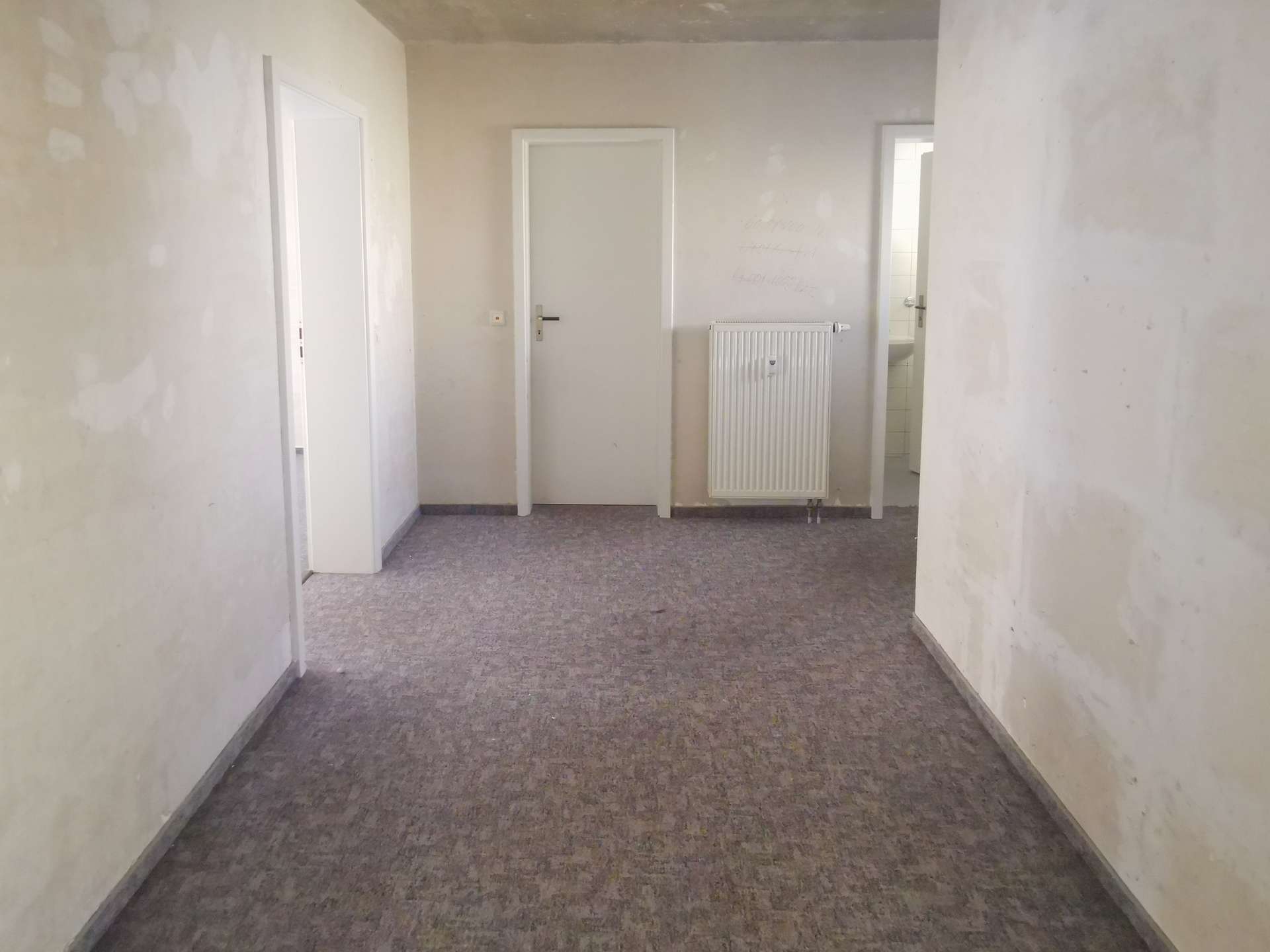 Unfinished indoor hallway with patched pale walls, a white door on the left, a radiator on the center wall, and an open doorway to a bright room at the back.