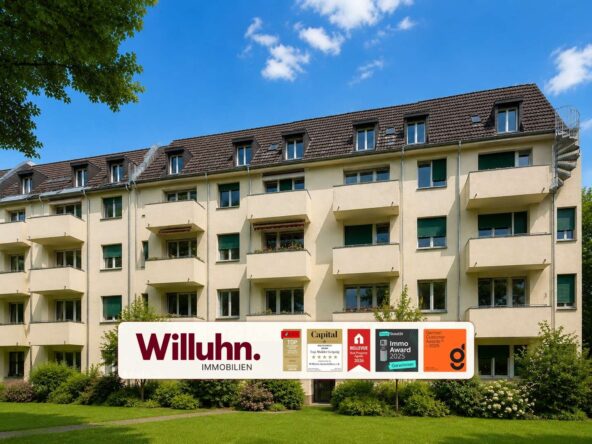 Beige residential building with multiple balconies under a blue sky; in front is a large Willuhn Immobilien sign with award plaques on the lawn.